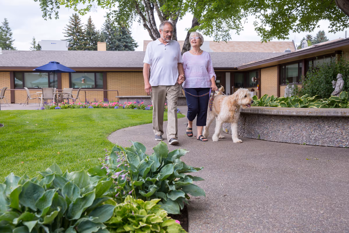 An older couple walk hand-in-hand with a large dog along a paved courtyard path in front of a low brick senior living building.