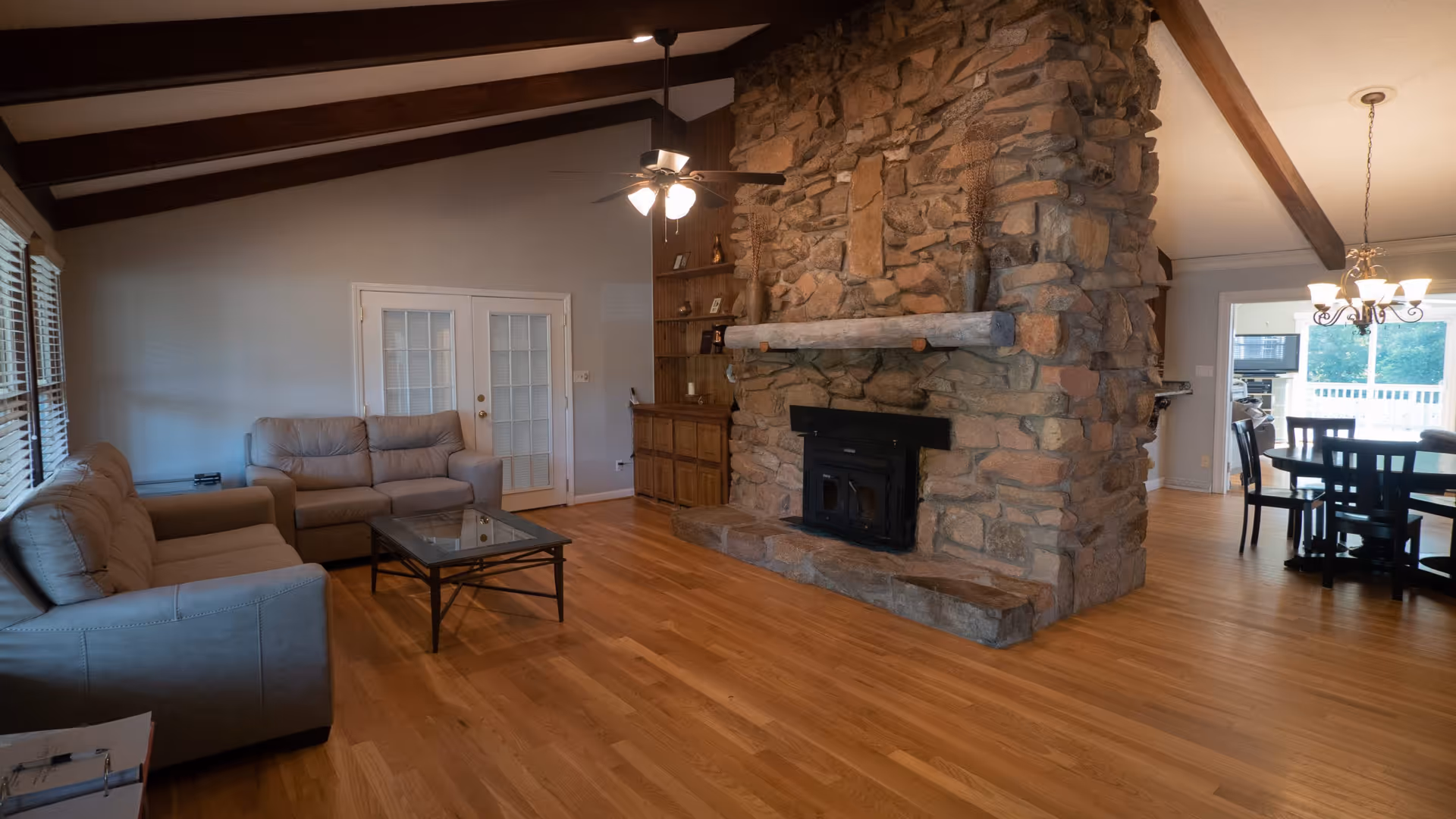 Spacious living room with vaulted beamed ceiling, large stone fireplace, sofas and a coffee table opening to a dining area.