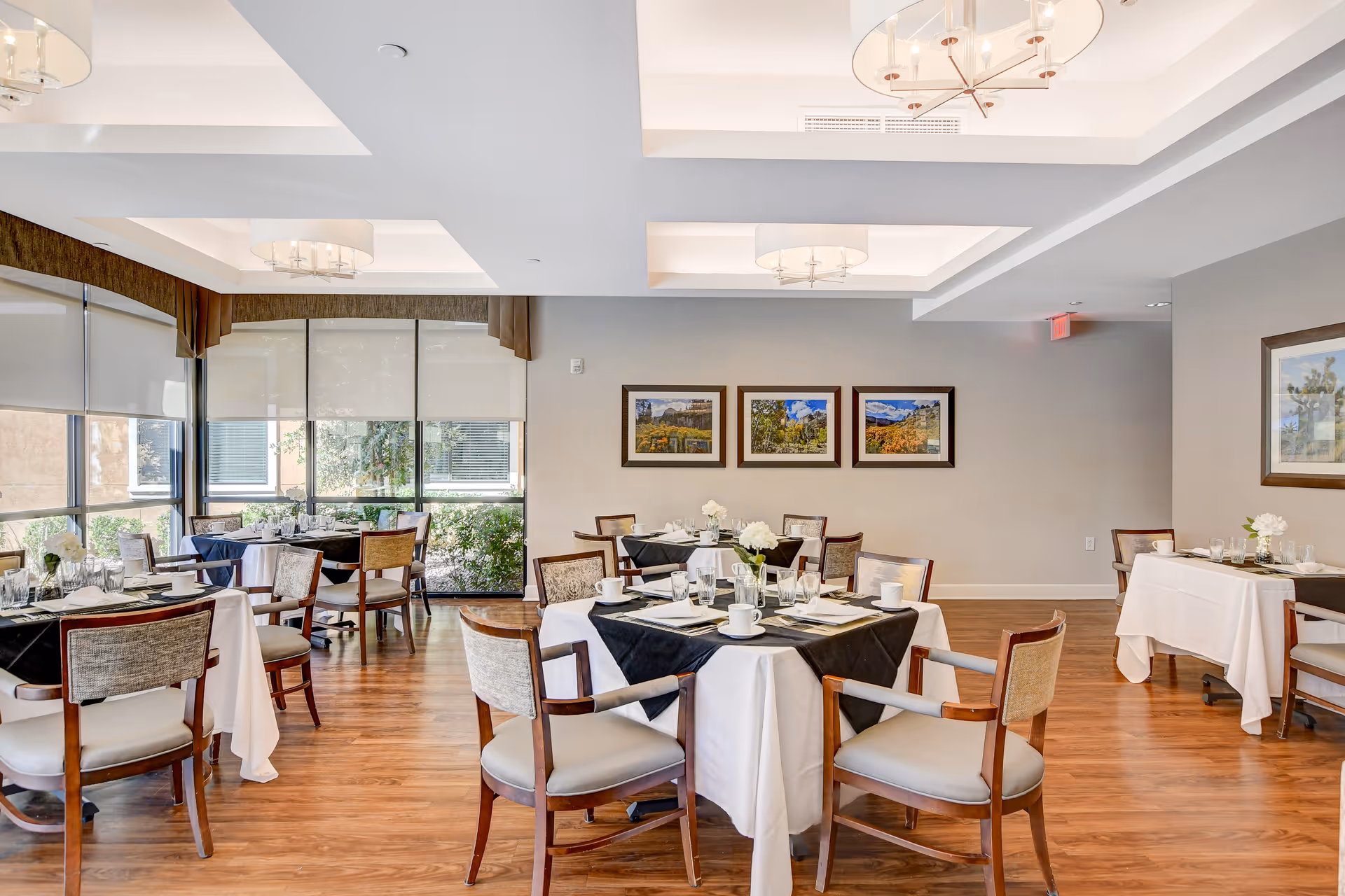 Bright dining room with multiple tables set for a meal, wooden chairs, large windows and framed artwork on the wall.
