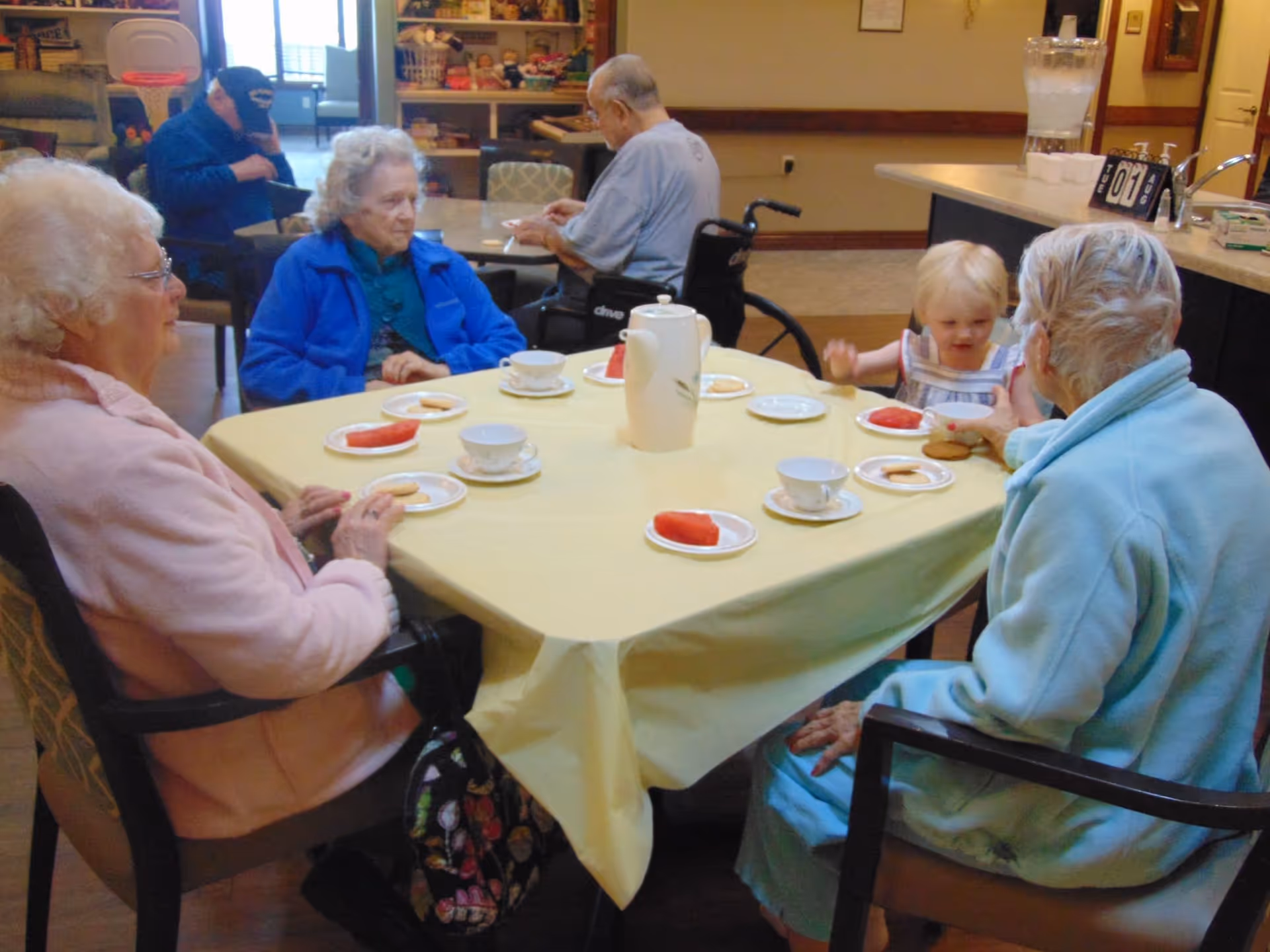 Several elderly residents and a small child sitting around a table with teacups and plates in a communal dining area.