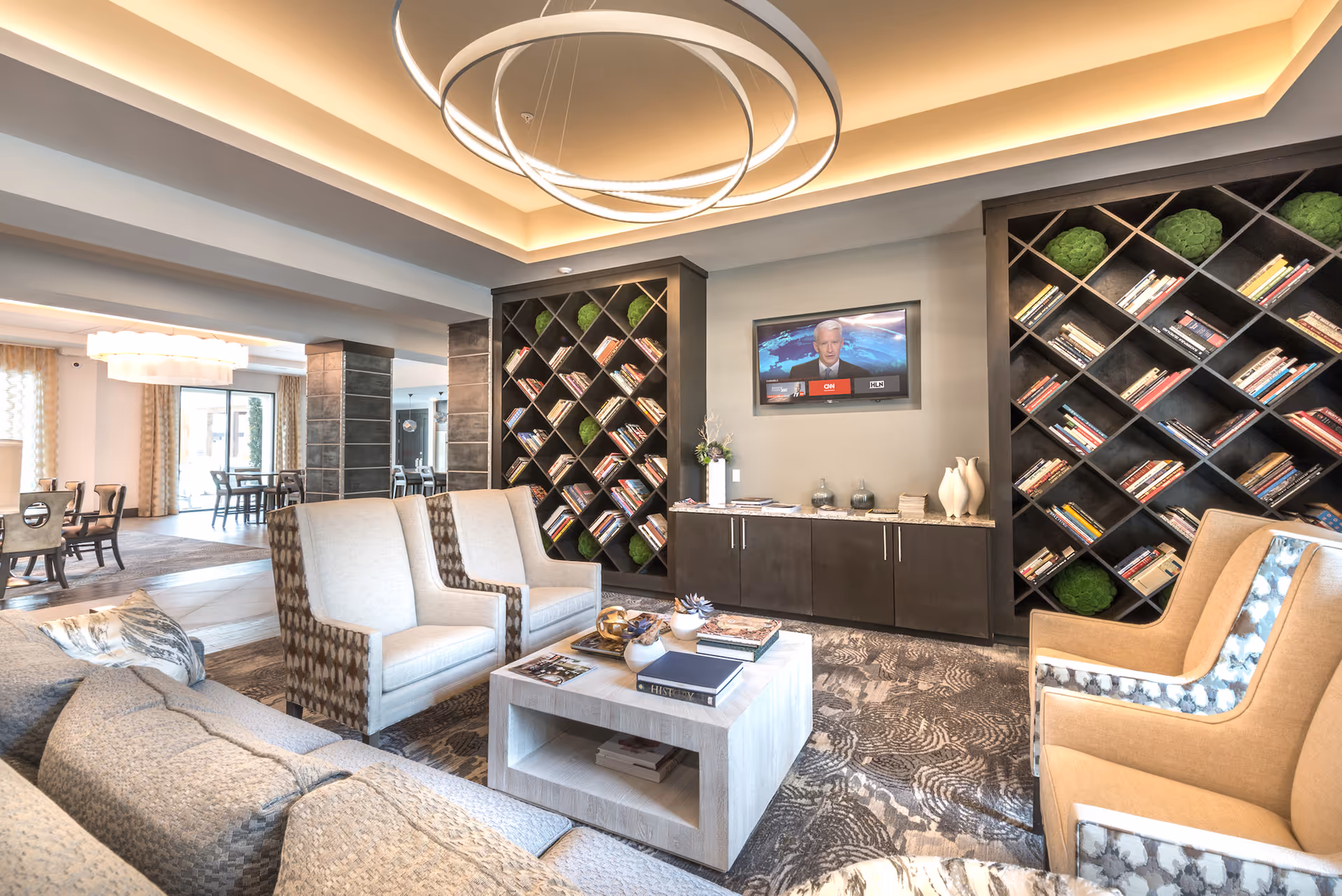 A modern and cozy living room area with comfortable armchairs and a sofa arranged around a light wooden coffee table. The room features built-in bookshelves with books and decorative green plants, a flat-screen TV mounted on the wall, and stylish ceiling lighting. In the background, there is a dining area with tables and chairs near large windows with curtains.