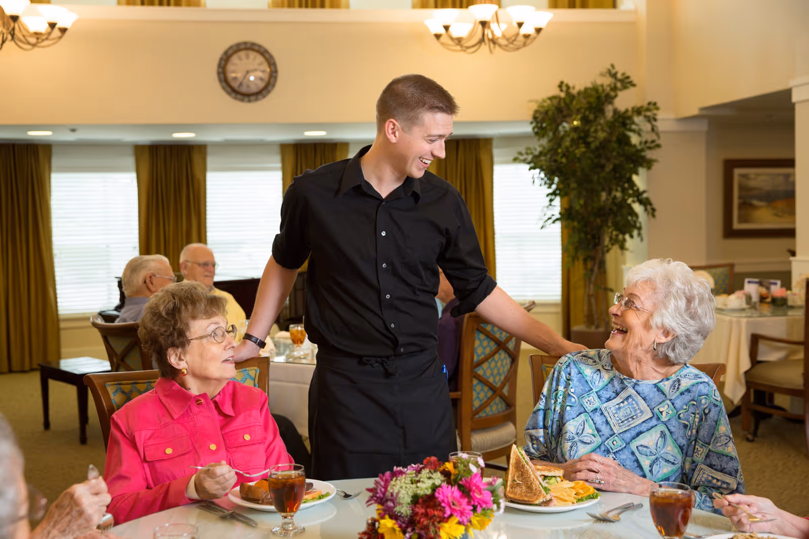 A young male server in a black uniform smiling and interacting with two elderly women seated at a dining table in a senior living facility. The women are enjoying their meals and drinks, with a bouquet of flowers on the table. Other elderly residents are visible in the background in a well-lit dining room with large windows and yellow curtains.