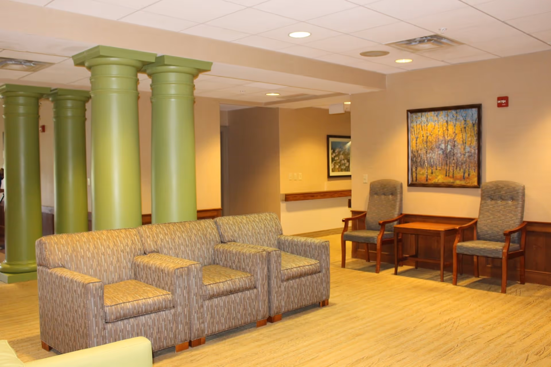 A seating area in a senior living facility with patterned armchairs and two wooden chairs with cushions around a small wooden table. The room features green columns, beige walls, a framed painting of a forest with autumn colors, and recessed ceiling lights.