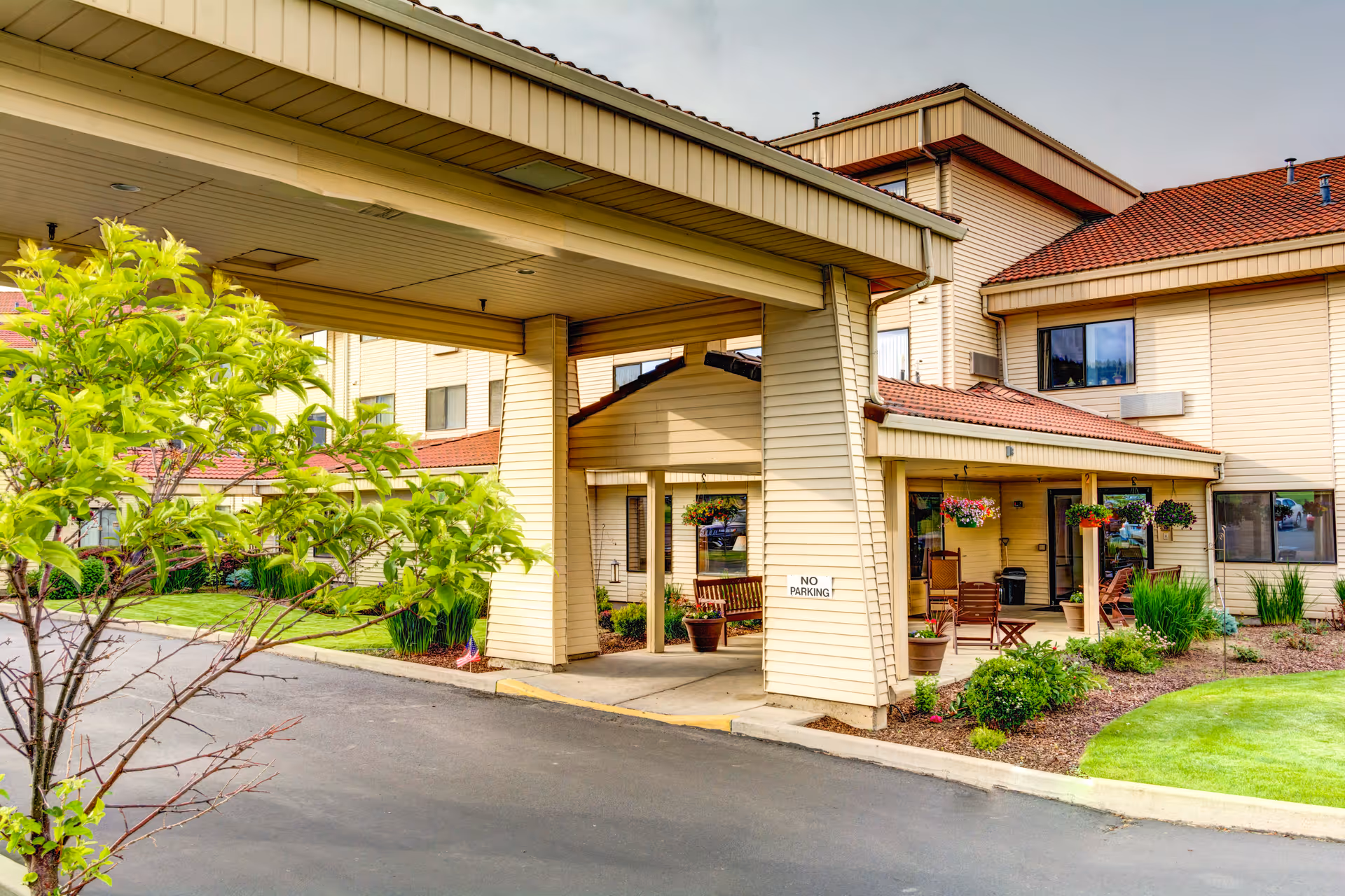 Exterior view of Grande Ronde Retirement Residence showing the entrance with a covered drop-off area, beige siding, red roof tiles, and a landscaped garden with green grass and plants. There are hanging flower pots and outdoor seating near the entrance.
