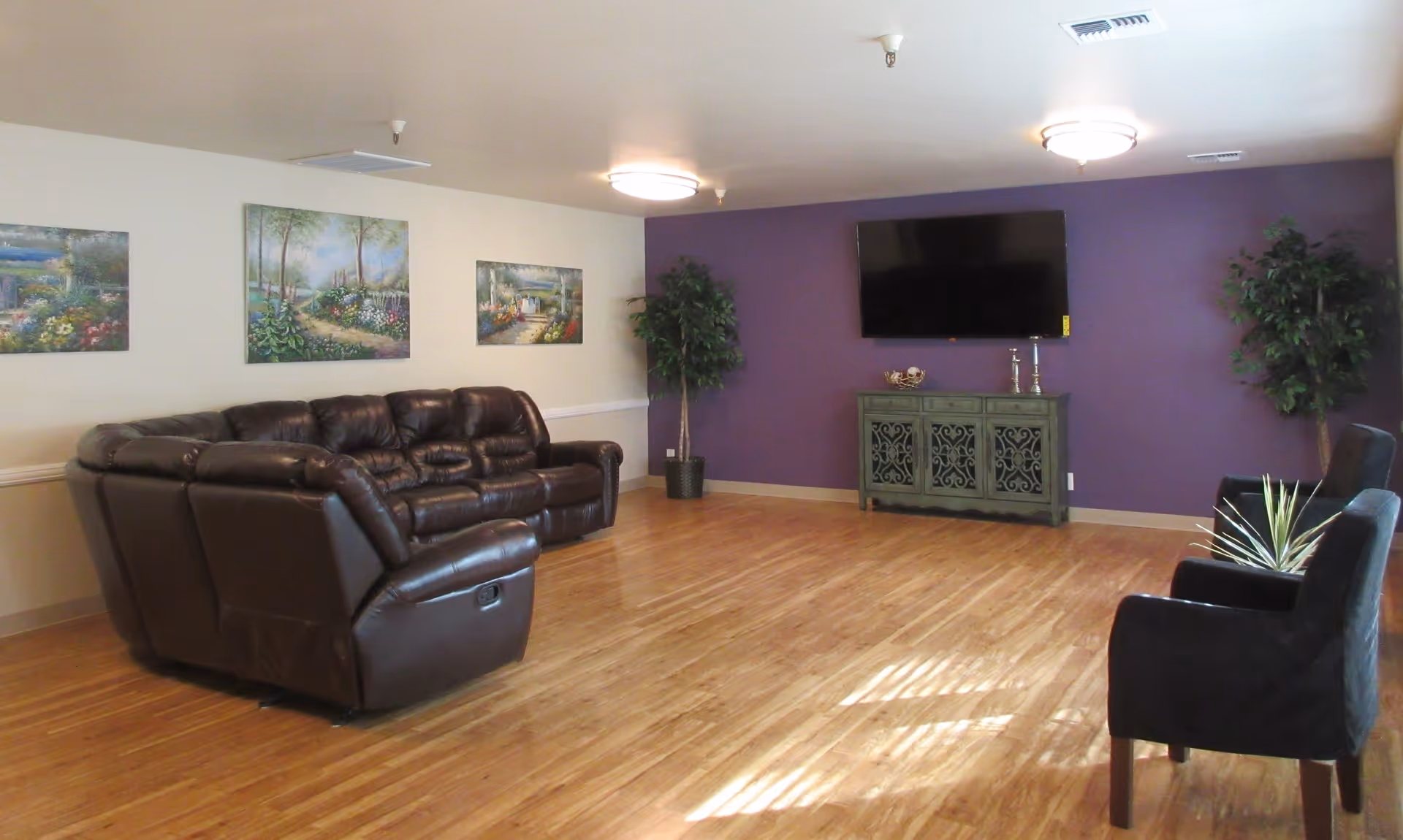 A spacious living room with a large brown leather sectional sofa on the left and two black armchairs on the right. The room features a wooden floor, a purple accent wall with a mounted flat-screen TV above a decorative cabinet, and three landscape paintings on the opposite wall. Two potted plants are placed near the TV wall, and the ceiling has two round light fixtures.
