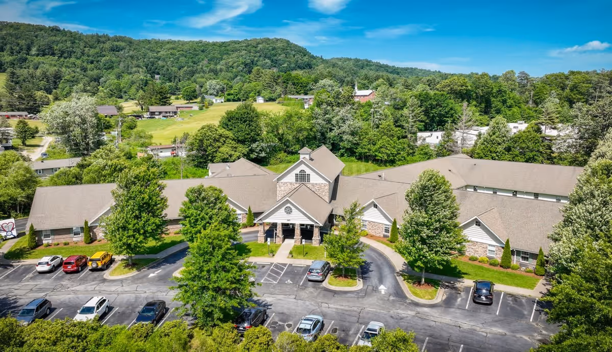 Aerial view of Deerfield Ridge Assisted Living facility surrounded by trees and greenery, with a parking lot in front and a backdrop of forested hills under a blue sky with some clouds.