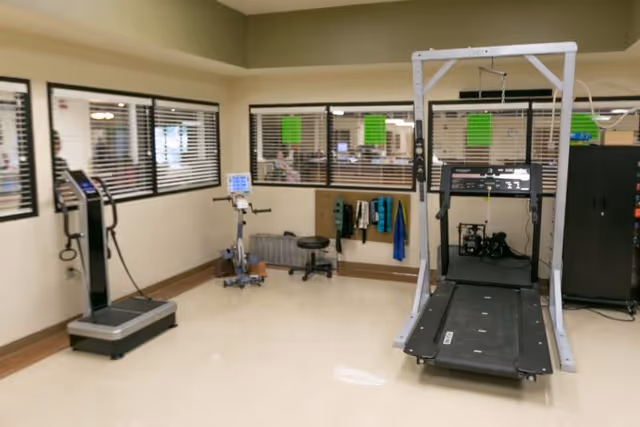 A rehabilitation room with exercise equipment including a treadmill, a stationary bike, and a weight scale. The room has beige walls, windows with blinds, and some towels hanging on a rack.