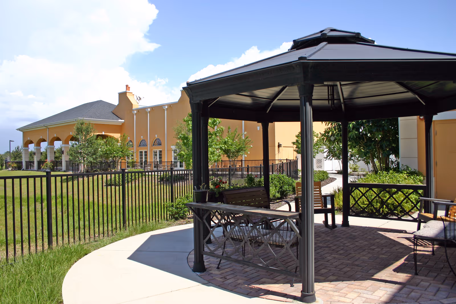 Outdoor gazebo with benches and chairs on a paved area surrounded by greenery and a black metal fence, with a large yellow building in the background under a partly cloudy blue sky.