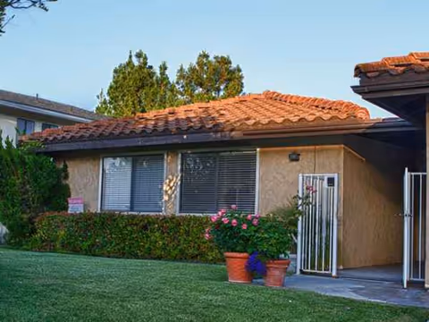 Single-story beige building with a red-tiled roof, hedge, lawn, and potted flowers.