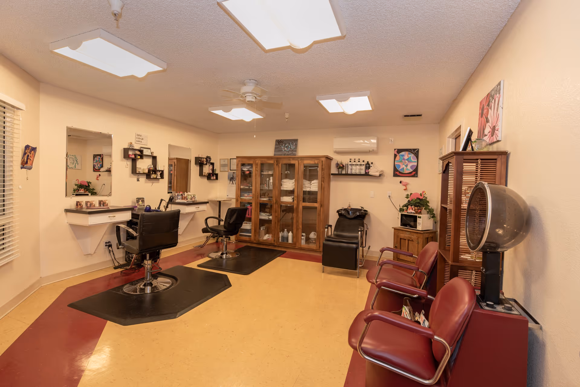 Small salon room with barber chairs, a shampoo sink, a hooded dryer, and cabinets in a senior living facility.