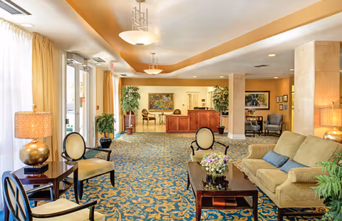 A spacious and well-lit senior living facility lobby with patterned blue and gold carpet, beige walls, and ceiling lights. The room features a seating area with a beige sofa, several chairs, a dark wooden coffee table with a floral arrangement, and side tables with lamps. There are large windows with sheer curtains on the left, potted plants, and a reception desk in the background.