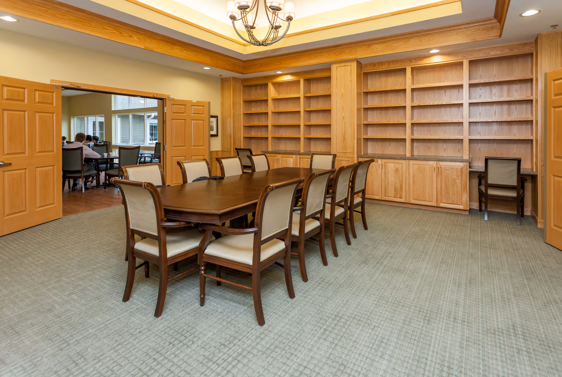 A well-lit room with a large wooden table surrounded by ten upholstered chairs. The room features built-in wooden shelves and cabinets along one wall, and double wooden doors open to an adjacent room where several people are seated at tables. The ceiling has a decorative light fixture and recessed lighting.
