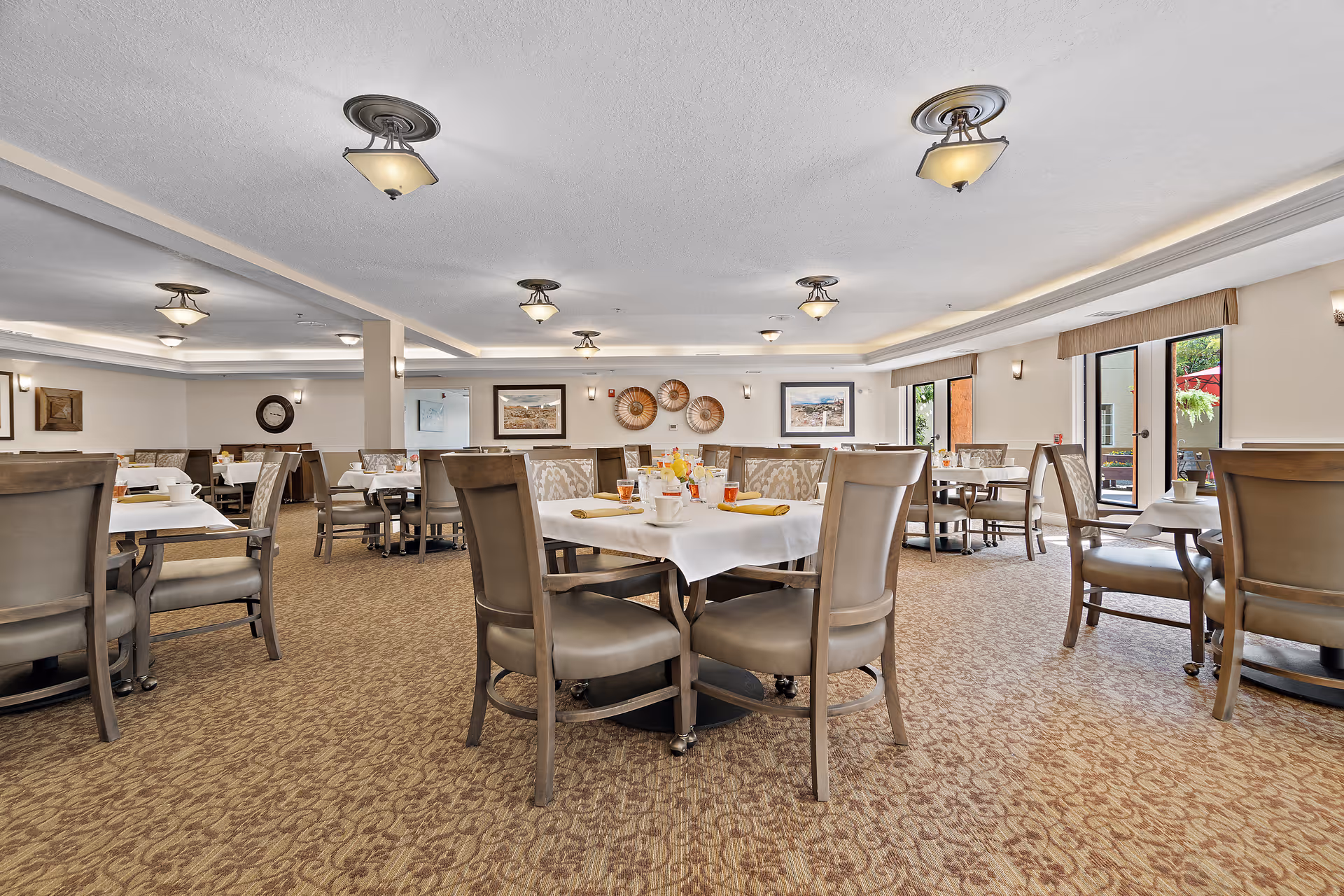 A spacious dining room with multiple tables and chairs arranged neatly. The tables are set with white tablecloths, cups, glasses, and napkins. The room has carpeted flooring, soft lighting from ceiling fixtures, and large windows letting in natural light. Wall decorations include framed pictures and woven baskets.