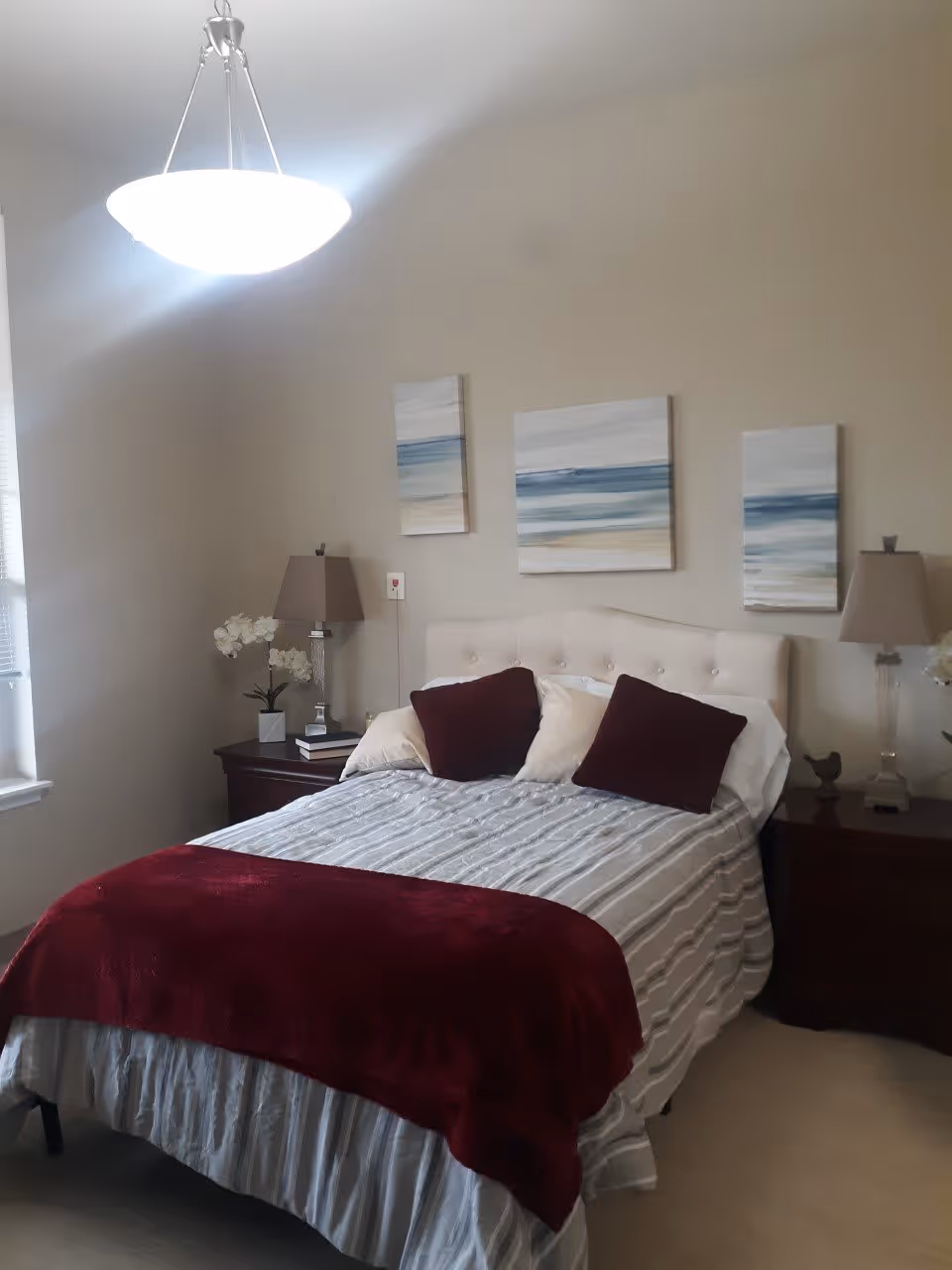 A neatly made bed with a white tufted headboard, striped bedding, and burgundy throw blanket and pillows. There are two matching bedside tables with lamps, a small plant, and decorative items. Three abstract paintings with blue and beige tones hang on the wall above the bed. A window with blinds is partially visible on the left side of the room, and a ceiling light fixture is above the bed.
