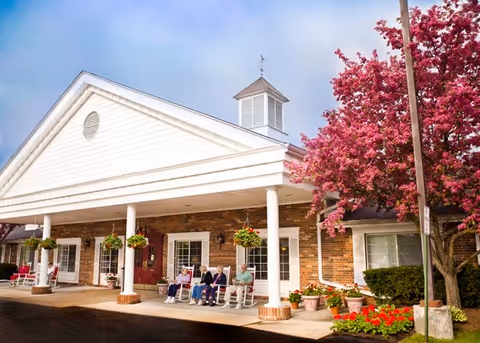 Front exterior view of a senior living facility with a white porch supported by columns, brick walls, hanging flower baskets, and a blooming pink tree on the right. Several elderly people are seated on rocking chairs on the porch.