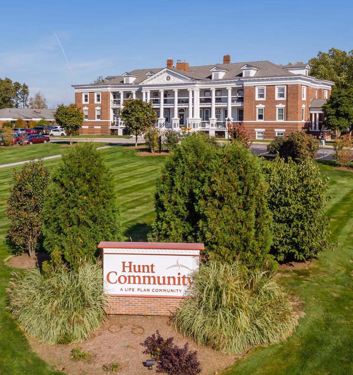 Exterior view of Hunt Community, a large brick building with white columns and a manicured lawn with trees and bushes in front. A sign in the foreground reads 'Hunt Community A Life Plan Community'.