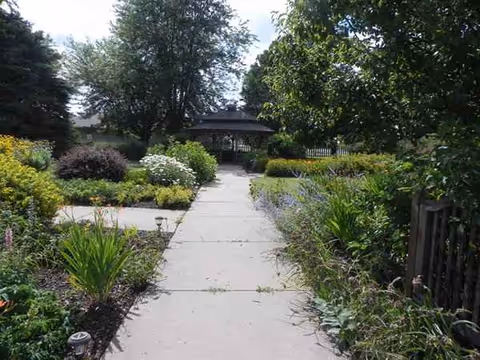 A garden path leads to a gazebo surrounded by trees and flower beds.