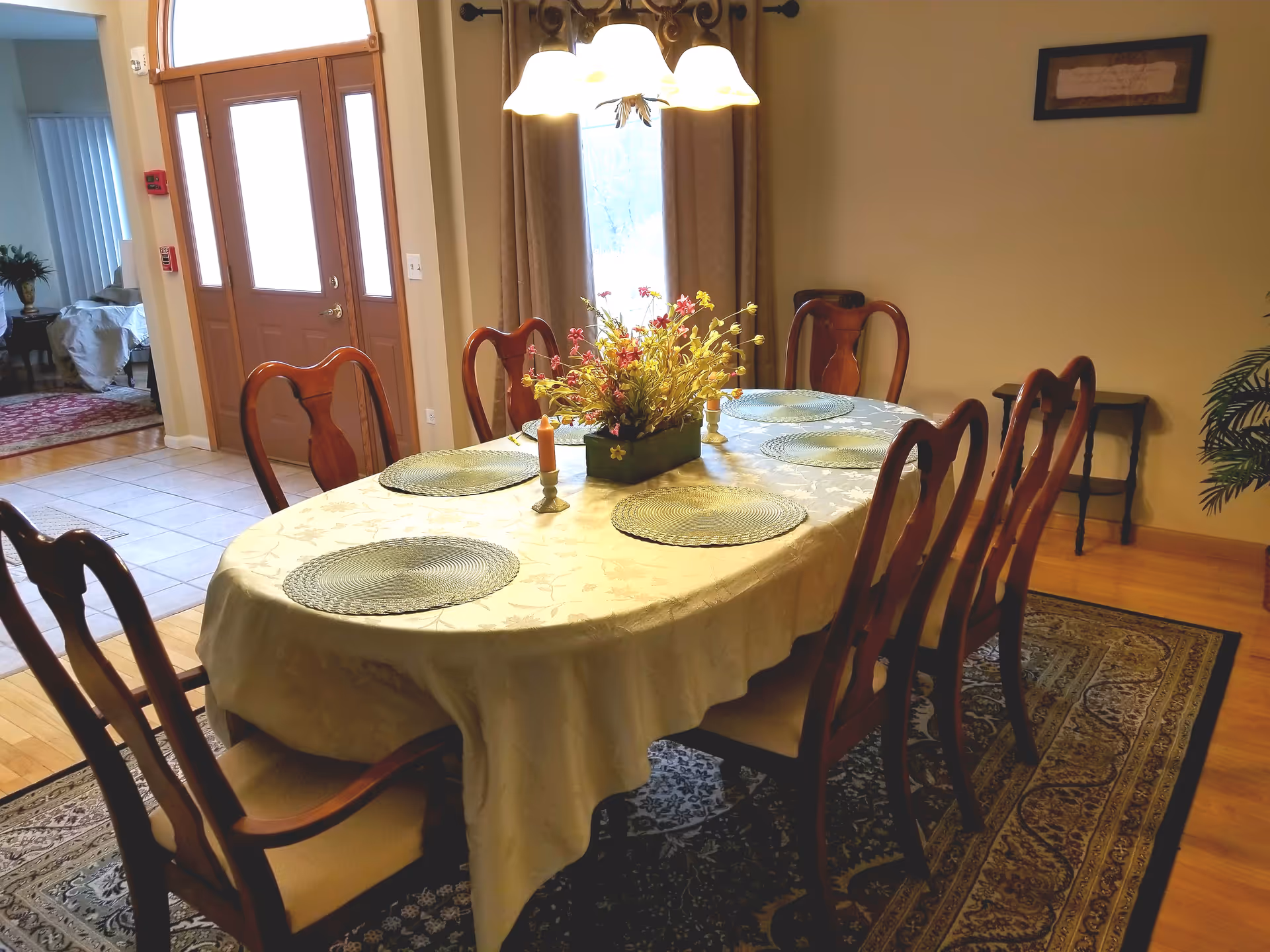A dining room with a wooden table covered by a cream-colored tablecloth, set with six round placemats and a floral centerpiece. Six wooden chairs with cushioned seats surround the table. A chandelier with five lights hangs above the table. In the background, there is a wooden front door with glass panels and a window with curtains. The floor is a combination of hardwood and tile, and a patterned rug is underneath the dining table.
