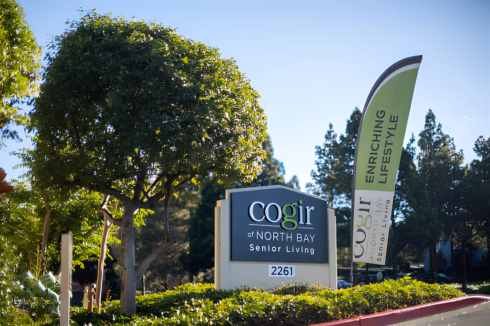 Outdoor view of the entrance sign for Cogir of North Bay Senior Living, surrounded by greenery and trees, with a tall green flag beside the sign that reads 'ENRICHING LIFESTYLE'.