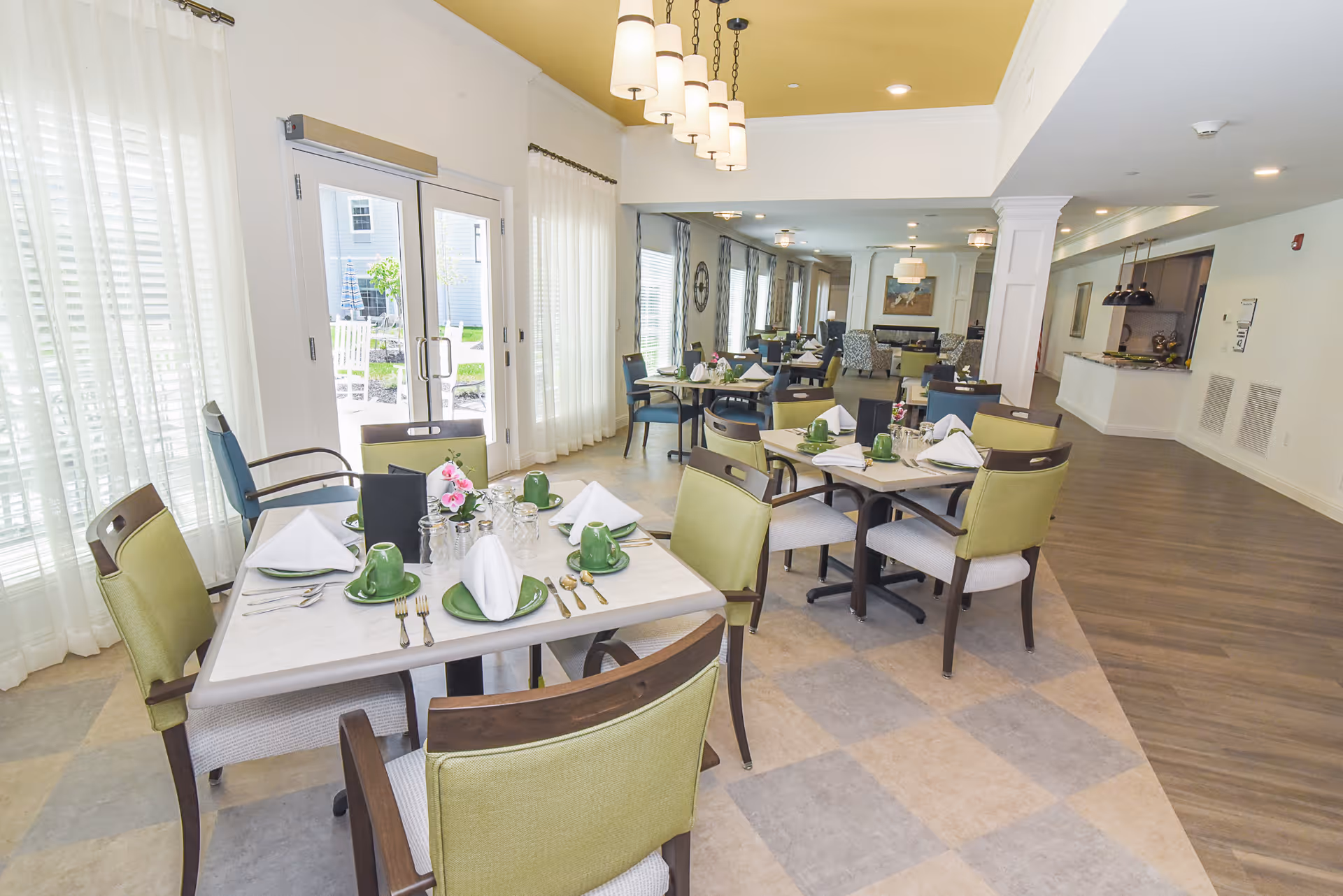 A bright dining room with several tables set for meals, each with green cups, plates, and neatly folded white napkins. The room has large windows with sheer white curtains allowing natural light to fill the space. There are green cushioned chairs around the tables, and modern pendant lights hang from the ceiling. In the background, there is a fireplace and additional seating areas.