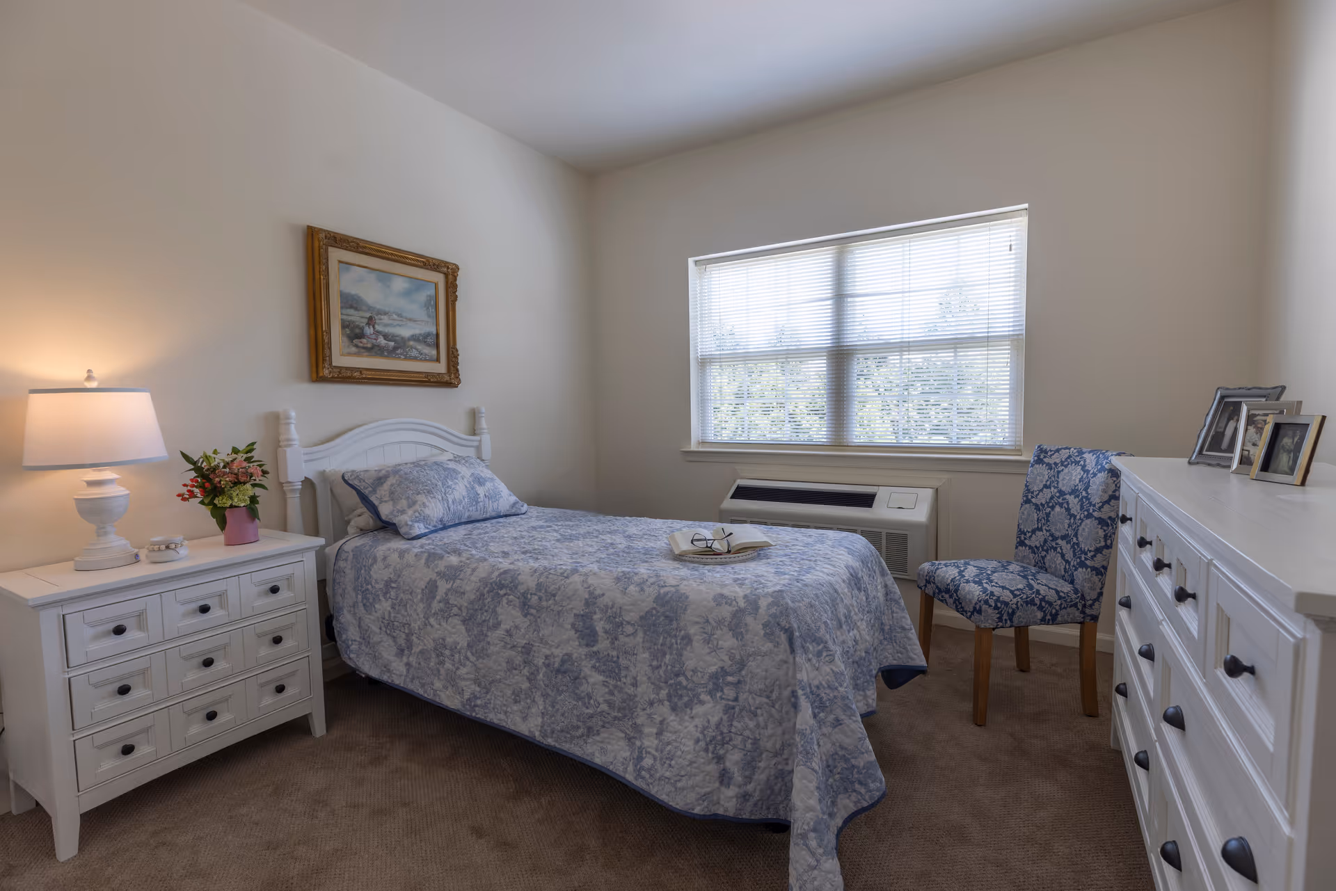 A cozy bedroom with a single bed covered in a blue and white patterned quilt. Next to the bed is a white nightstand with a lamp and a vase of flowers. A framed painting hangs on the wall above the bed. Across from the bed is a white dresser with framed photos on top and a blue patterned chair beside it. A window with blinds lets in natural light.