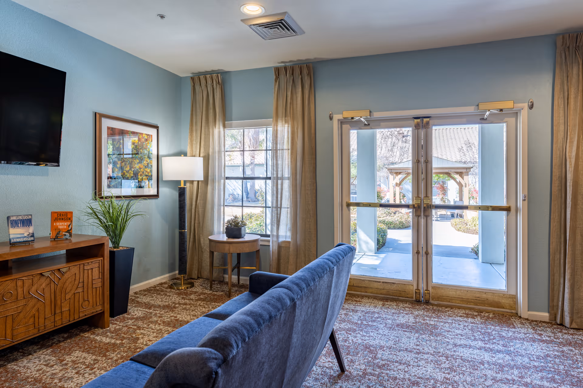 A cozy living room area with a blue sofa facing a wooden cabinet with books and a potted plant. A floor lamp stands next to a window with beige curtains, and double glass doors lead outside to a garden area with a gazebo.