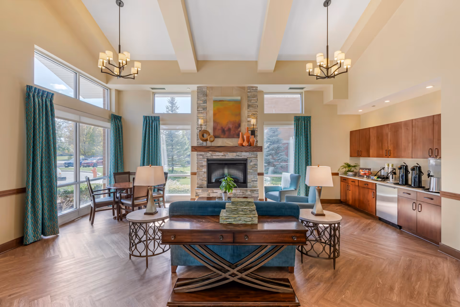 Sunlit senior living common room with seating around a stone fireplace, large windows, and a kitchenette area.
