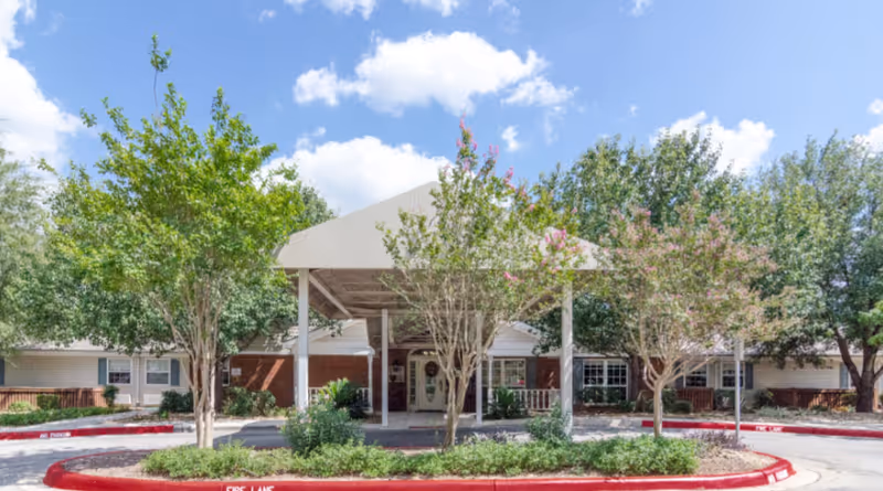 Covered main entrance of a single-story brick senior living facility with a circular driveway, landscaping, and trees under a blue sky.