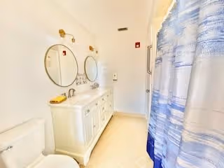 Bright bathroom with white walls and floor, featuring a white vanity with two round mirrors above it, gold light fixtures, a toilet, and a shower with a blue and white striped curtain.