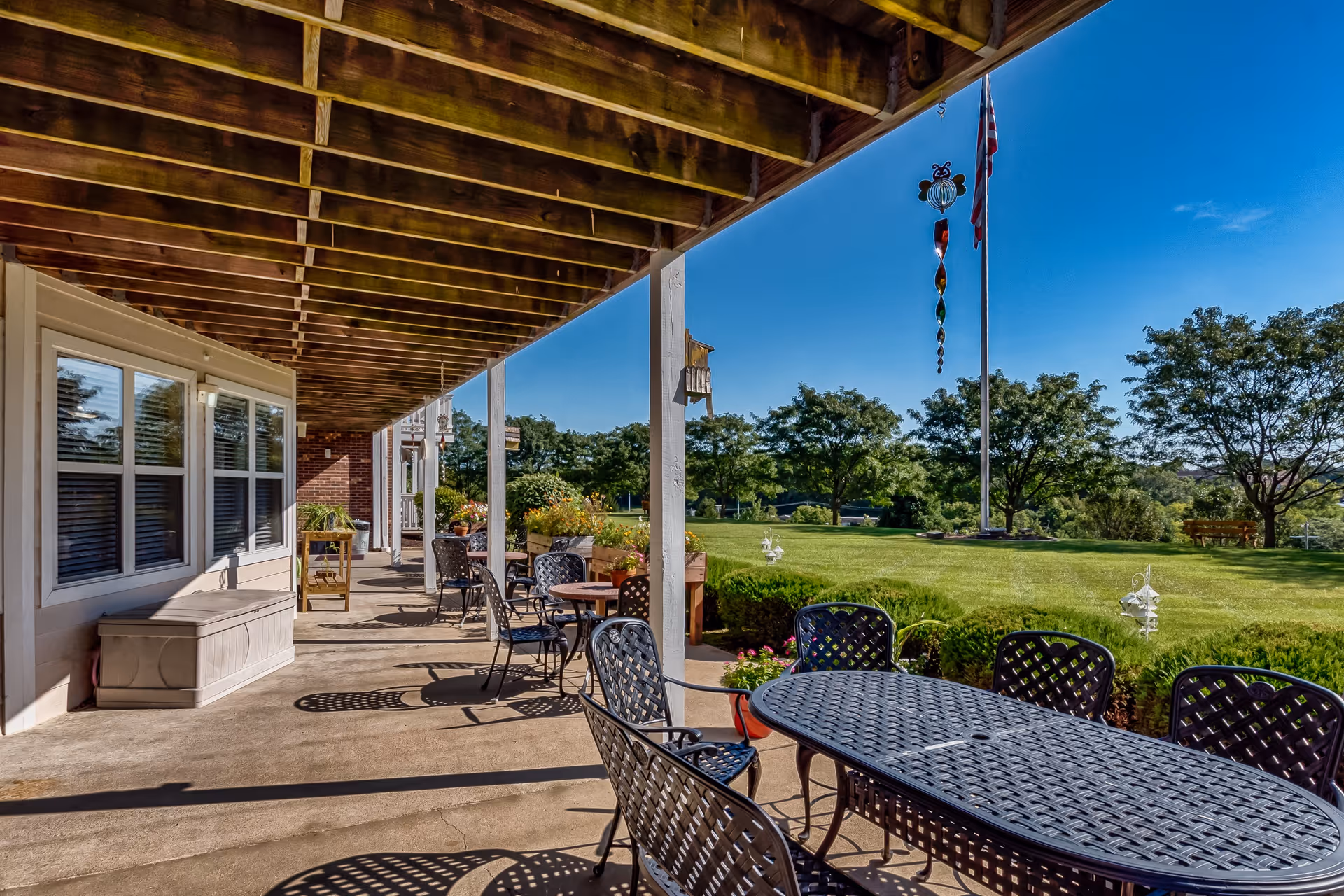 Covered outdoor patio area with metal tables and chairs, overlooking a well-maintained grassy lawn with trees and a flagpole under a clear blue sky.