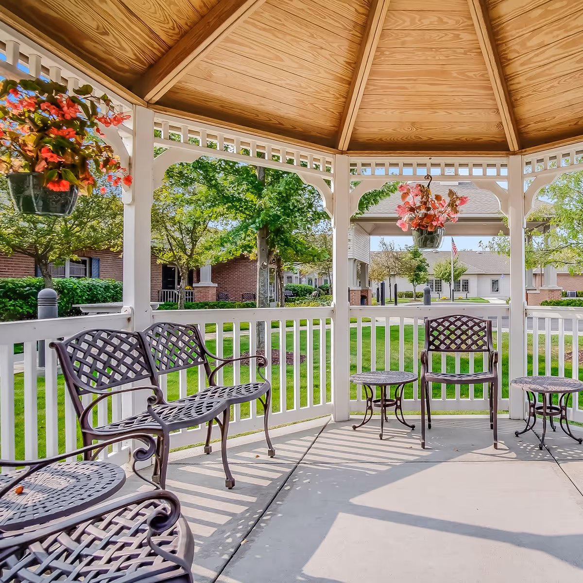View from inside a wooden gazebo with a pitched roof, featuring metal chairs and small tables. Hanging flower pots with red and pink flowers are attached to the gazebo. Outside the gazebo, there is a well-maintained green lawn, trees, and buildings in the background under a clear sky.