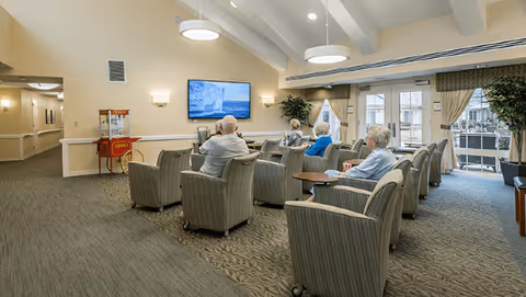 Several elderly residents sit in upholstered armchairs facing a wall-mounted TV in a bright communal lounge with large windows.