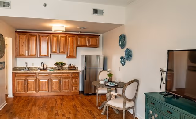 Interior view of a senior living facility kitchen and dining area featuring wooden cabinets, a stainless steel refrigerator, a small round dining table set with two chairs, decorative wall art, and a television on a green cabinet.