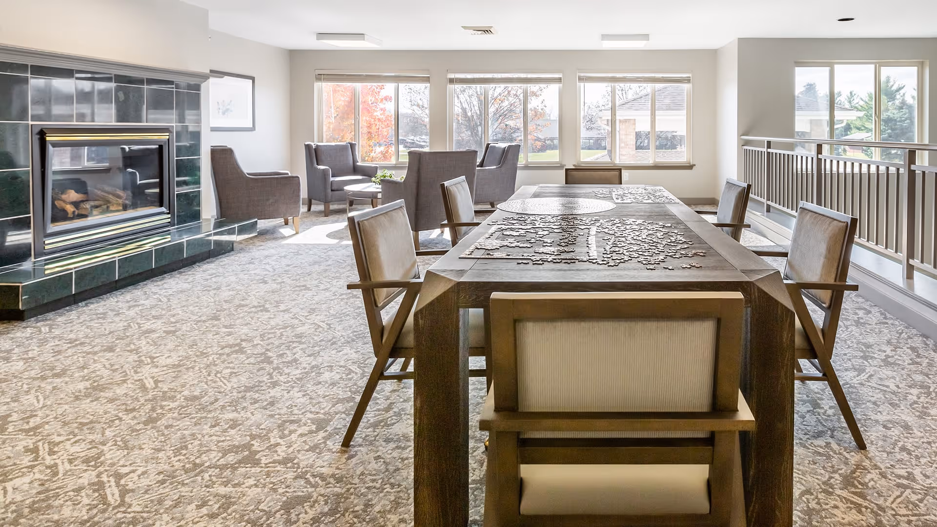 A bright and spacious common area with a large wooden table in the center covered with a partially completed jigsaw puzzle. Surrounding the table are six cushioned chairs. In the background, there are four armchairs arranged near large windows that let in natural light. To the left, there is a fireplace with a dark green tiled surround. The room has light-colored walls and carpeted flooring.