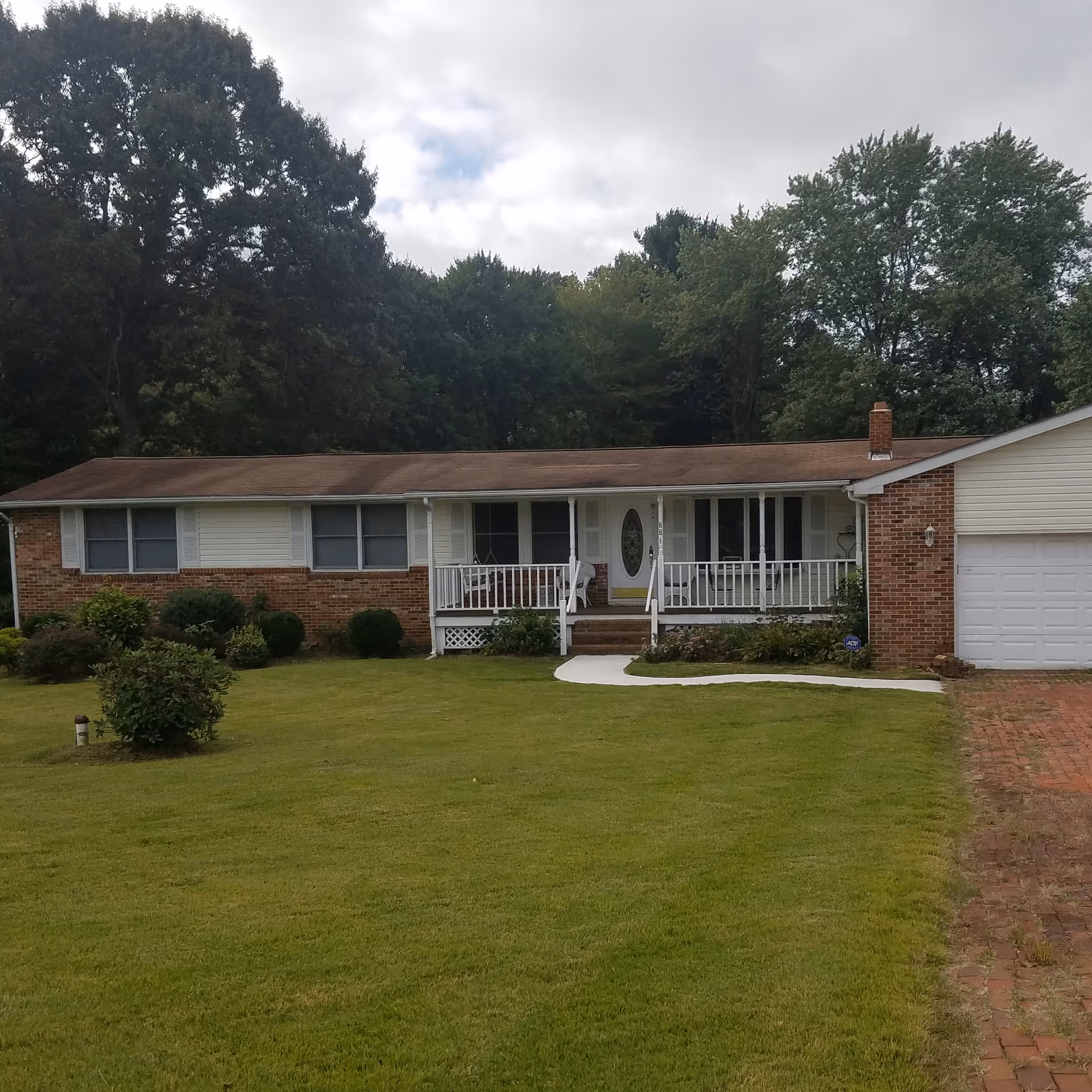 Single-story brick and siding house with a front porch, white railing, and a well-maintained lawn. There are several bushes and trees surrounding the house, and a driveway leading to a white garage door on the right side.