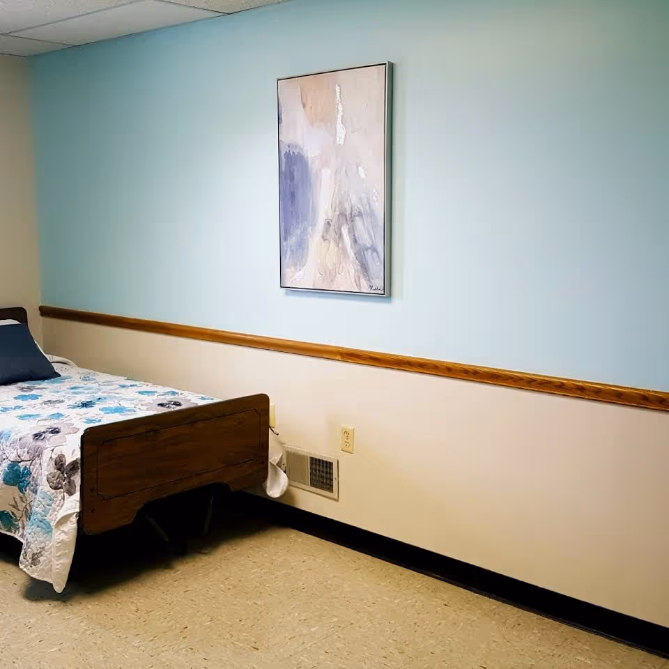 A simple bedroom with a single wooden bed featuring a floral patterned bedspread and a dark pillow. The walls are painted light blue and beige with a wooden trim separating the colors. A framed abstract painting with soft pastel colors hangs on the wall above the bed. The floor is covered with light-colored linoleum tiles.