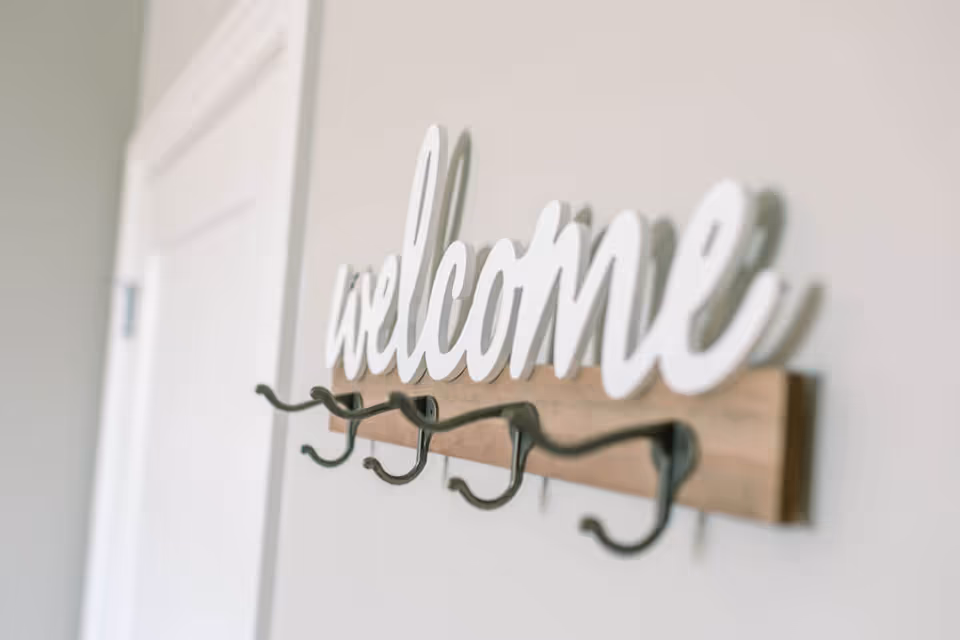 Wall-mounted wooden coat rack with a decorative white 'welcome' sign and several metal hooks next to a door.