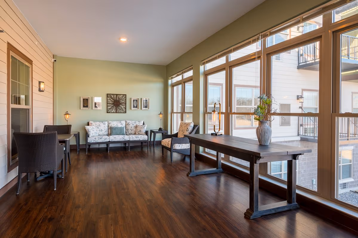 Bright communal lounge with a sofa, chairs, a long wooden table and large windows along one wall.
