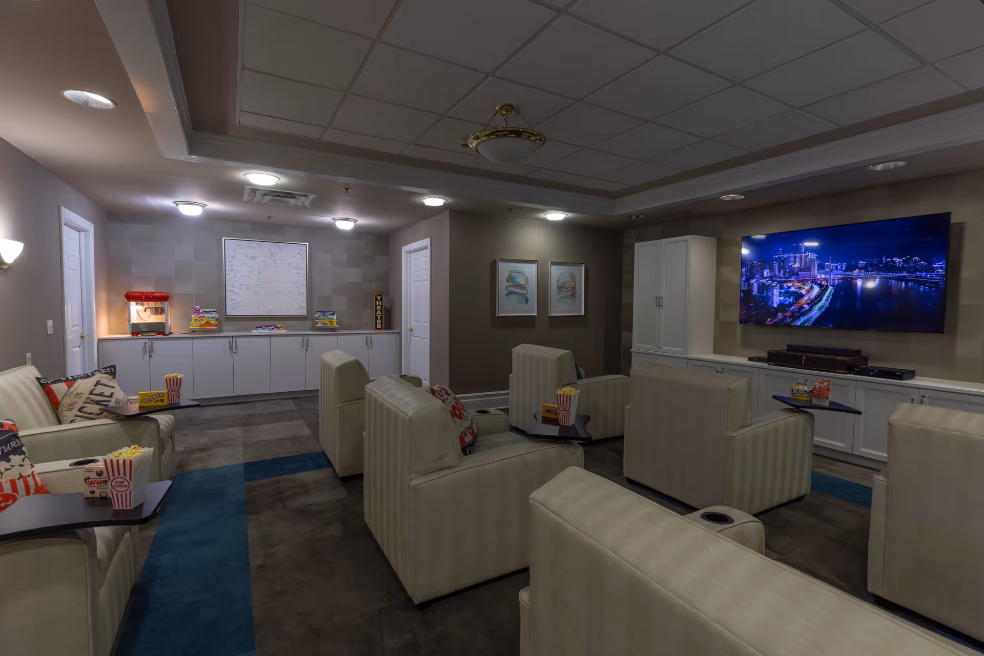A cozy media room with several beige armchairs arranged in rows facing a large flat-screen TV mounted on the wall. The TV displays a nighttime cityscape. There are small tables attached to some chairs holding popcorn containers. The room has a popcorn machine and snacks on a counter along the back wall, with soft lighting and framed artwork on the walls.