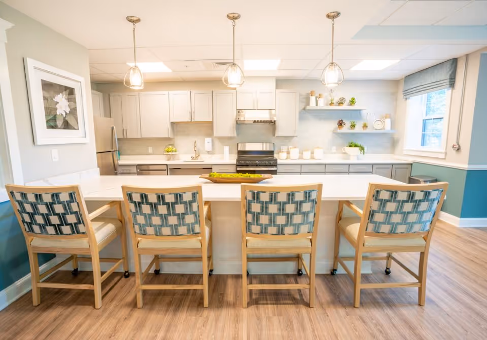 Bright and modern kitchen with a large white island countertop surrounded by four wooden chairs with patterned cushions. The kitchen features light gray cabinets, stainless steel appliances including a refrigerator and stove, pendant lights hanging above the island, and decorative plants on shelves and counters. There is a window with a blue roman shade on the right side and a framed flower picture on the left wall.