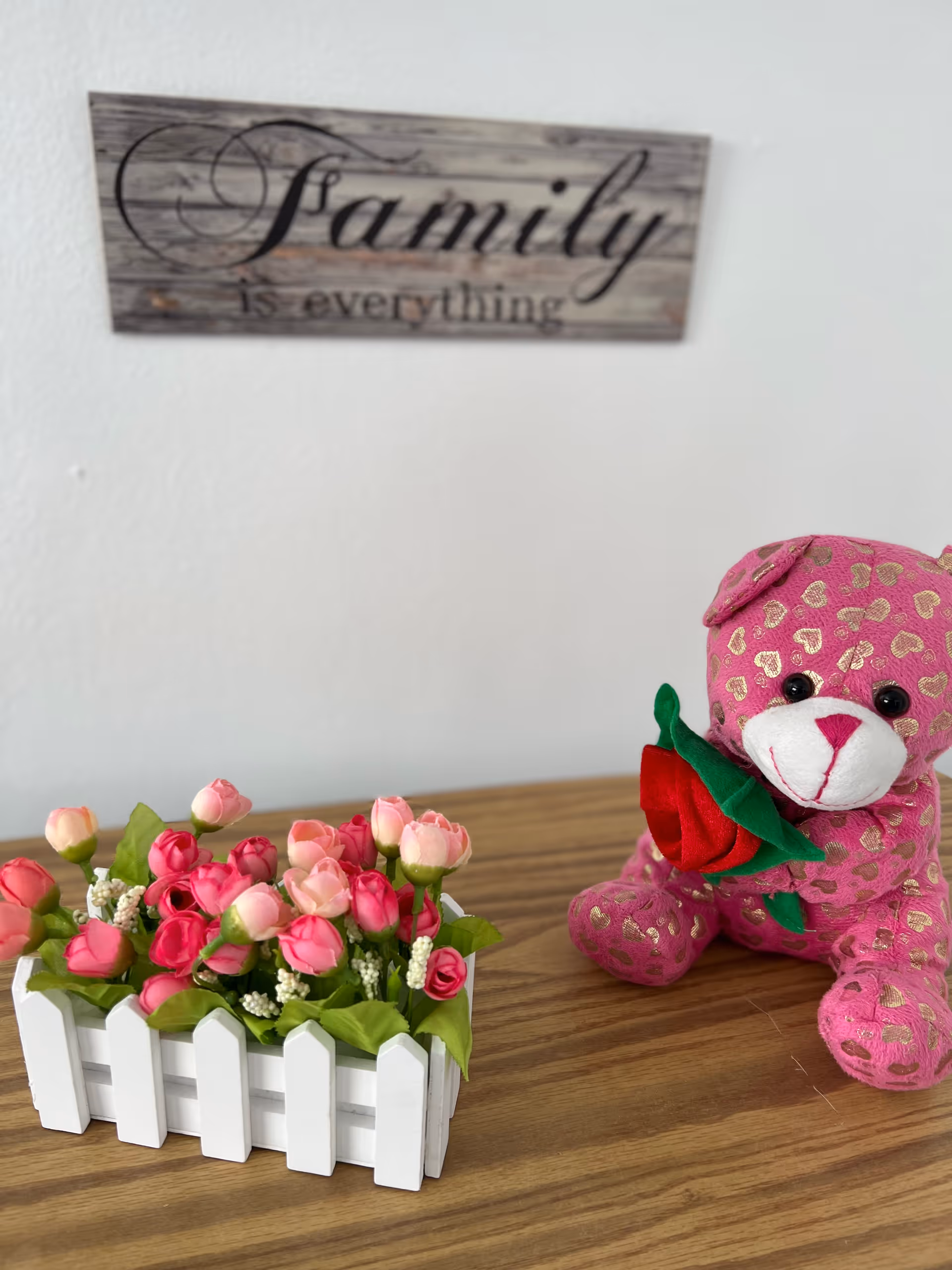 A wooden table with a small white picket fence planter holding pink and red artificial flowers, next to a pink stuffed teddy bear with gold heart patterns holding a red fabric rose. On the wall behind, there is a wooden sign with the text 'Family is everything'.