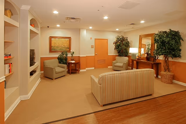 A cozy communal living room with a striped sofa, armchairs, built-in shelving, potted plants, and warm overhead lighting.