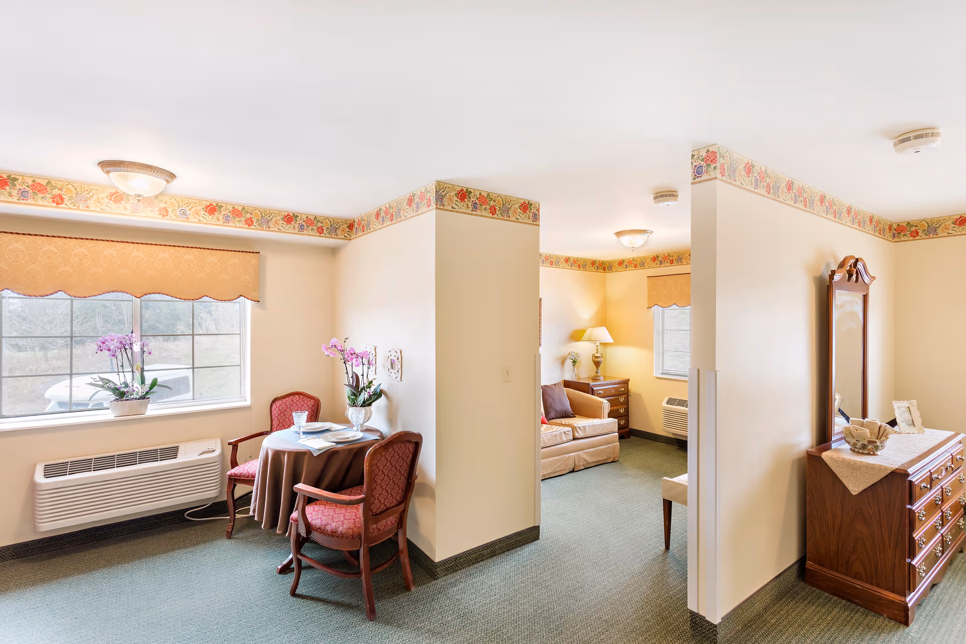 Interior view of a retirement village suite with a small dining area featuring a round table covered with a brown tablecloth, two red upholstered chairs, and a vase with purple flowers. Adjacent to the dining area is a living space with a beige sofa, a wooden side table with a lamp, and a window with a beige valance. A wooden dresser with a mirror and decorative items is visible on the right side. The walls are cream-colored with floral wallpaper borders near the ceiling, and the floor is carpeted in green.