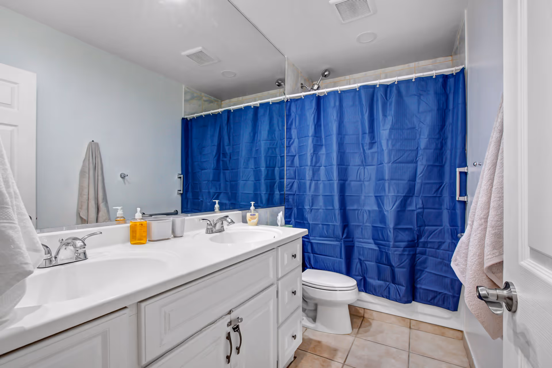 A clean bathroom with a white double sink vanity, two faucets, a large mirror above the sink, a toilet, and a bathtub with a blue shower curtain. There are towels hanging on the wall and door, and soap dispensers on the sink counter.
