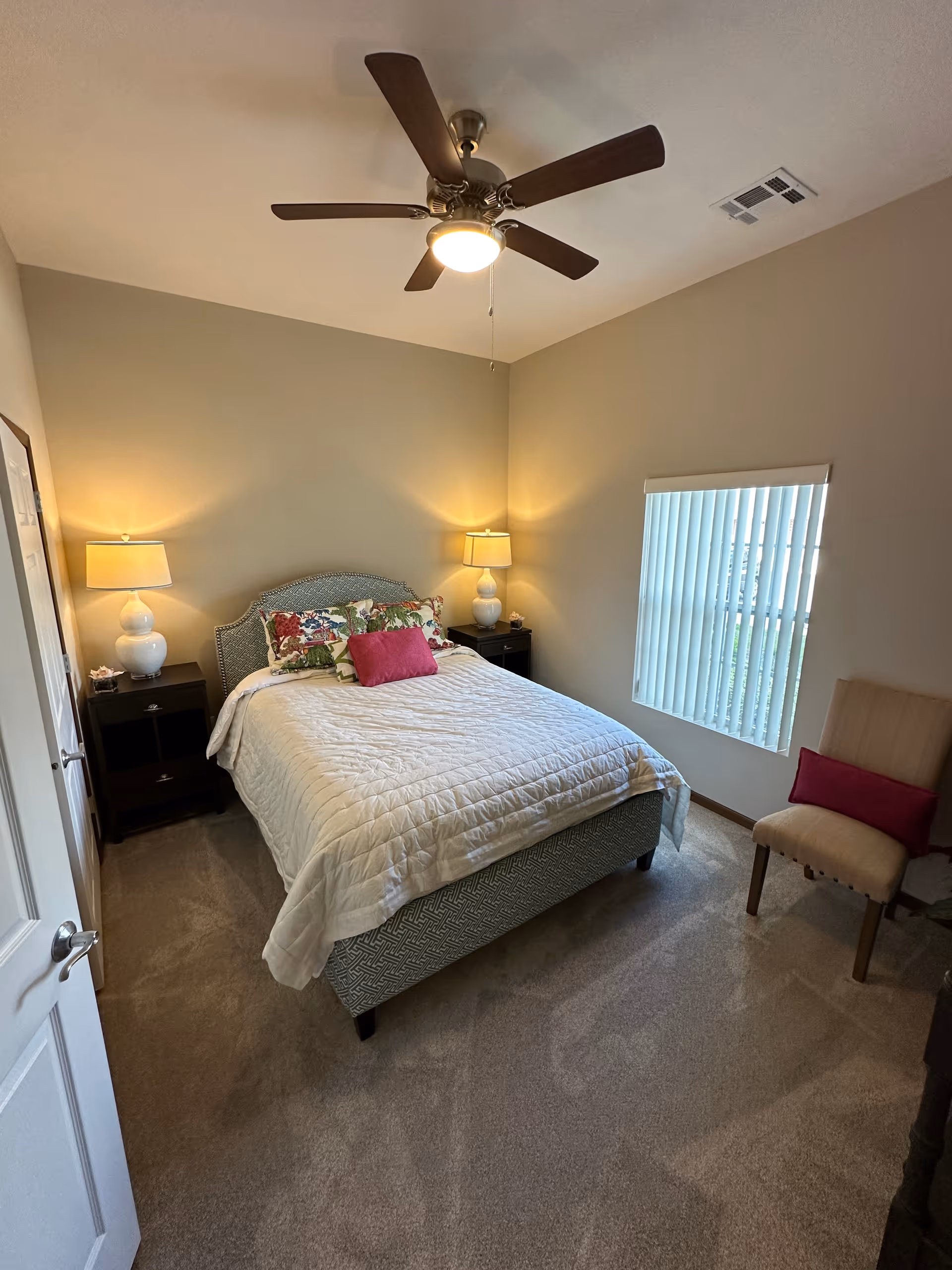 Neat bedroom with a bed, two bedside tables and lamps, ceiling fan, window blinds, and a chair.
