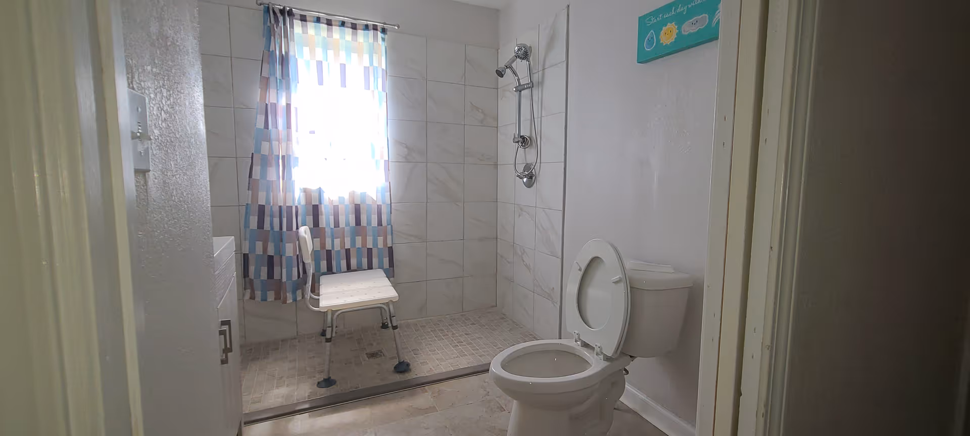 A bathroom with a walk-in shower featuring a shower chair and a window with a colorful curtain. There is a toilet with the lid open and a small cabinet visible on the left side. A decorative sign is hung on the wall above the toilet.