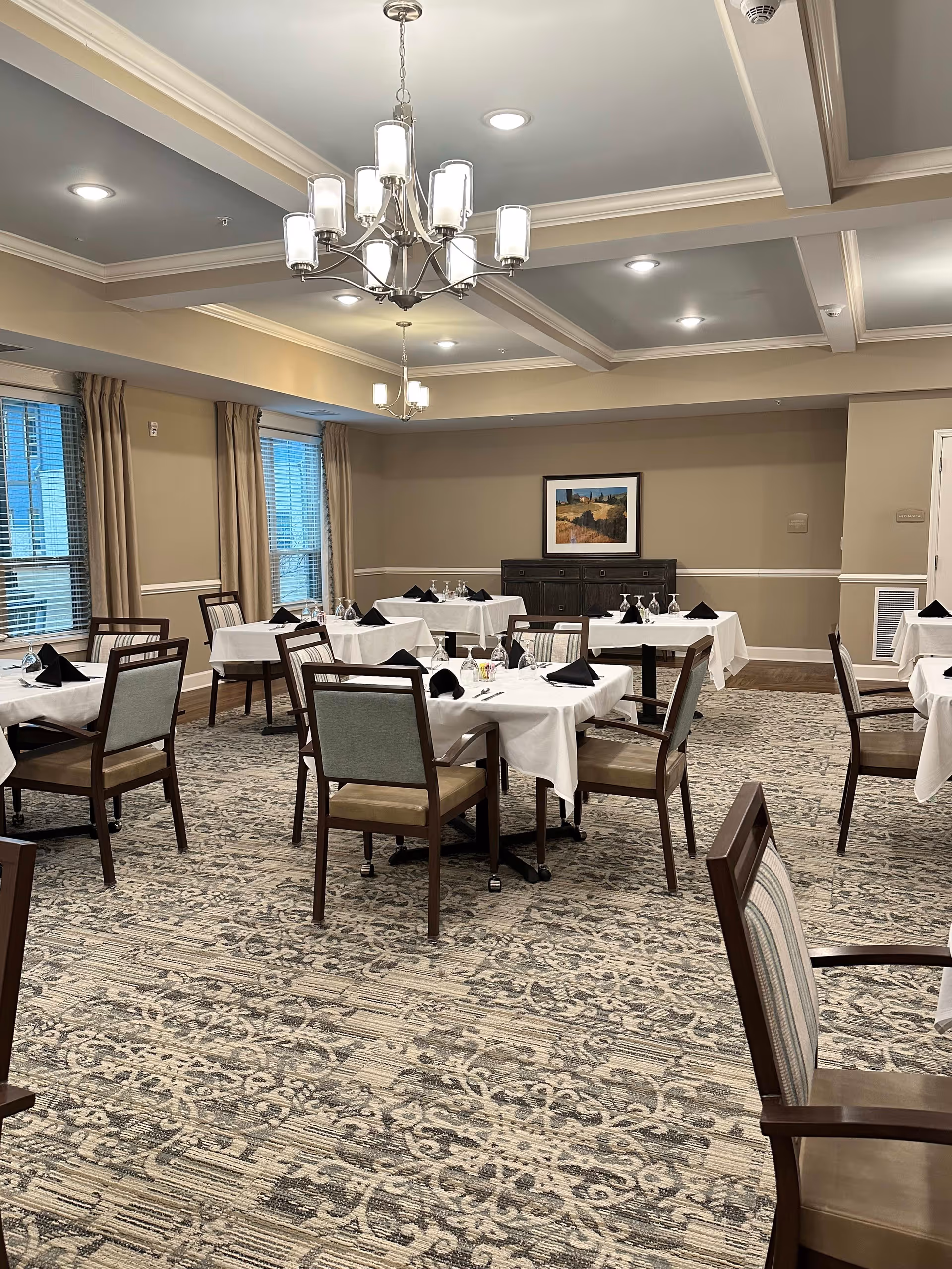 A dining room with several tables covered in white tablecloths, each set with black folded napkins and glassware. The room has beige walls, large windows with beige curtains, a patterned carpet, and a chandelier hanging from the ceiling. A framed landscape painting is on the far wall above a dark wooden sideboard.