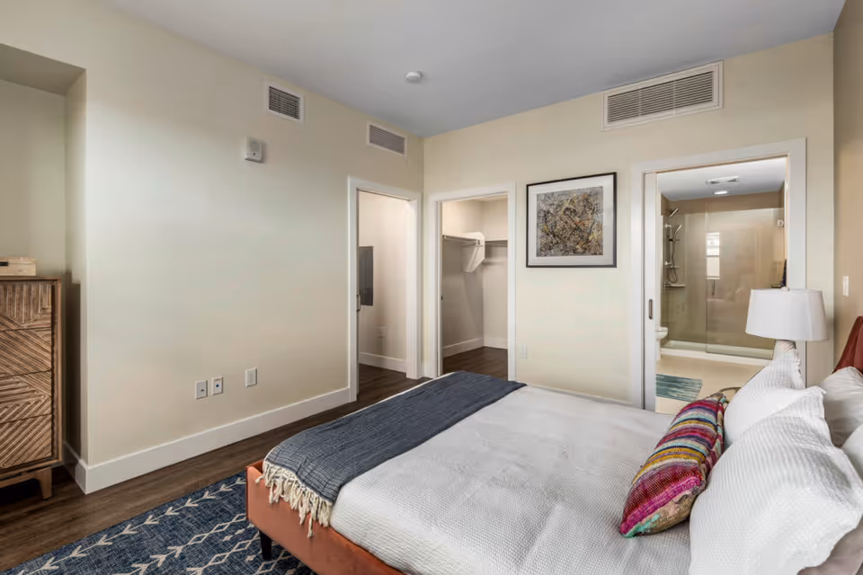 A bedroom with a neatly made bed featuring white bedding, a colorful pillow, and a gray throw blanket. The room has light-colored walls, a wooden floor, and a blue patterned rug. There is a wooden dresser on the left side. Two doorways are visible: one leading to a walk-in closet and the other to a bathroom with a glass-enclosed shower. A framed abstract artwork hangs on the wall between the two doorways.