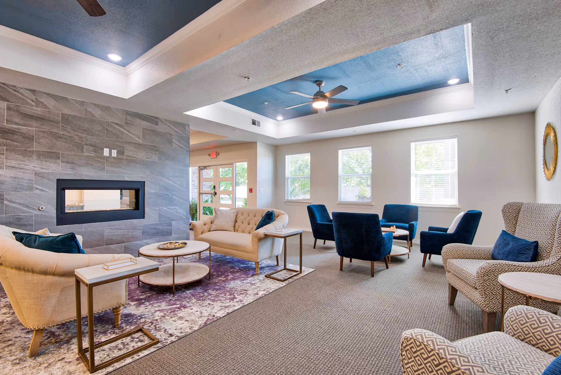 A bright and spacious living room in The Carlstone Independent 55 Living facility featuring a modern fireplace set in a tiled wall, beige and blue upholstered sofas and armchairs arranged around circular coffee tables, a blue accent ceiling with recessed lighting and ceiling fans, and large windows letting in natural light.