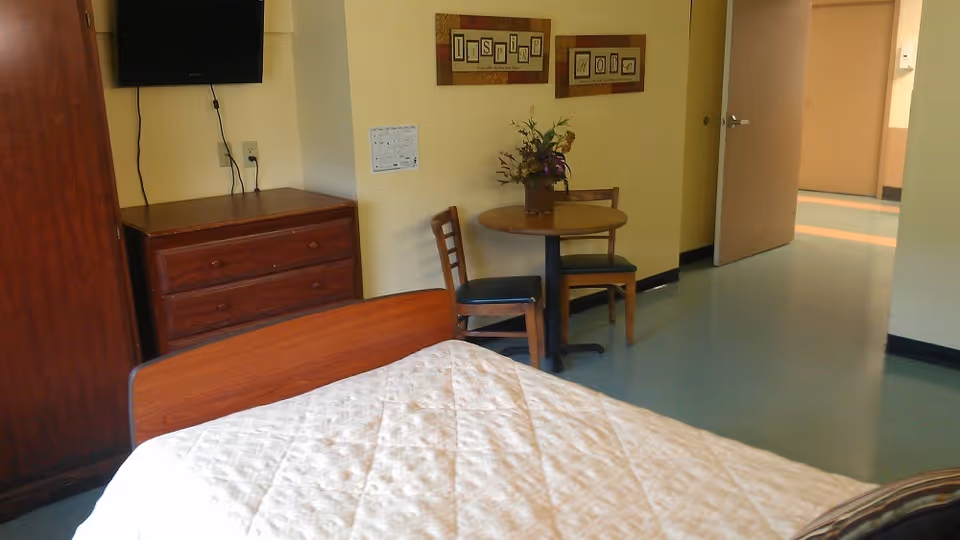 A nursing facility room with a bed covered in a white quilted blanket in the foreground. There is a wooden dresser with a flat-screen TV mounted above it on the wall. Next to the dresser is a small round table with two wooden chairs and a vase with flowers on top. The walls are painted light yellow, and there is a framed artwork with the words 'Inspire 2012' hanging above the table. An open door leads to a hallway with light blue flooring.